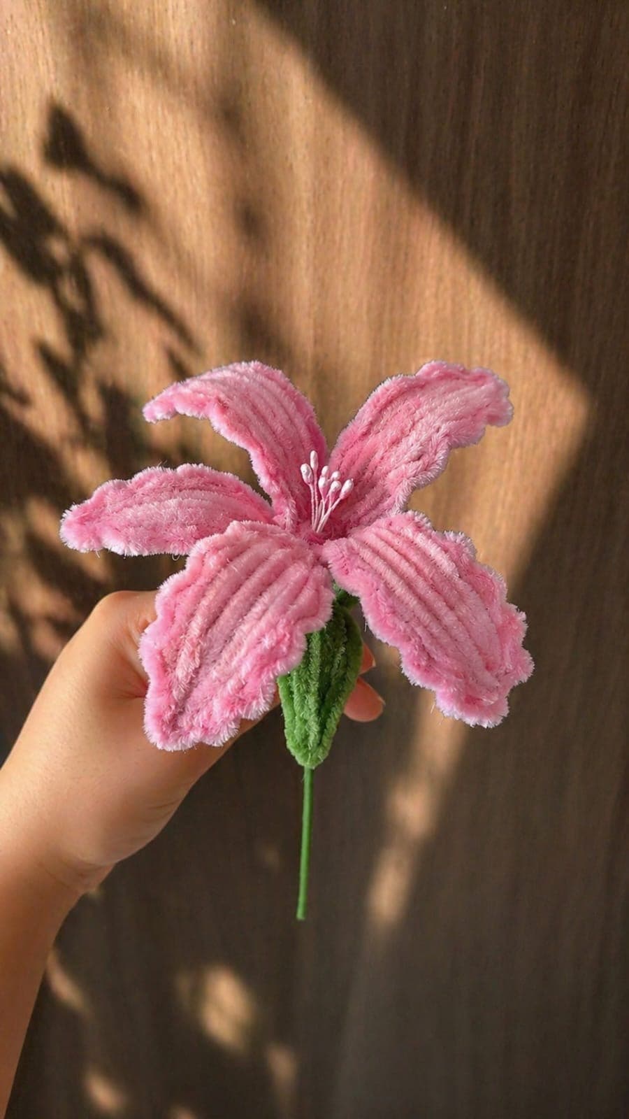 A plush rose-pink crochet lily with a beaded centre, held in hand.